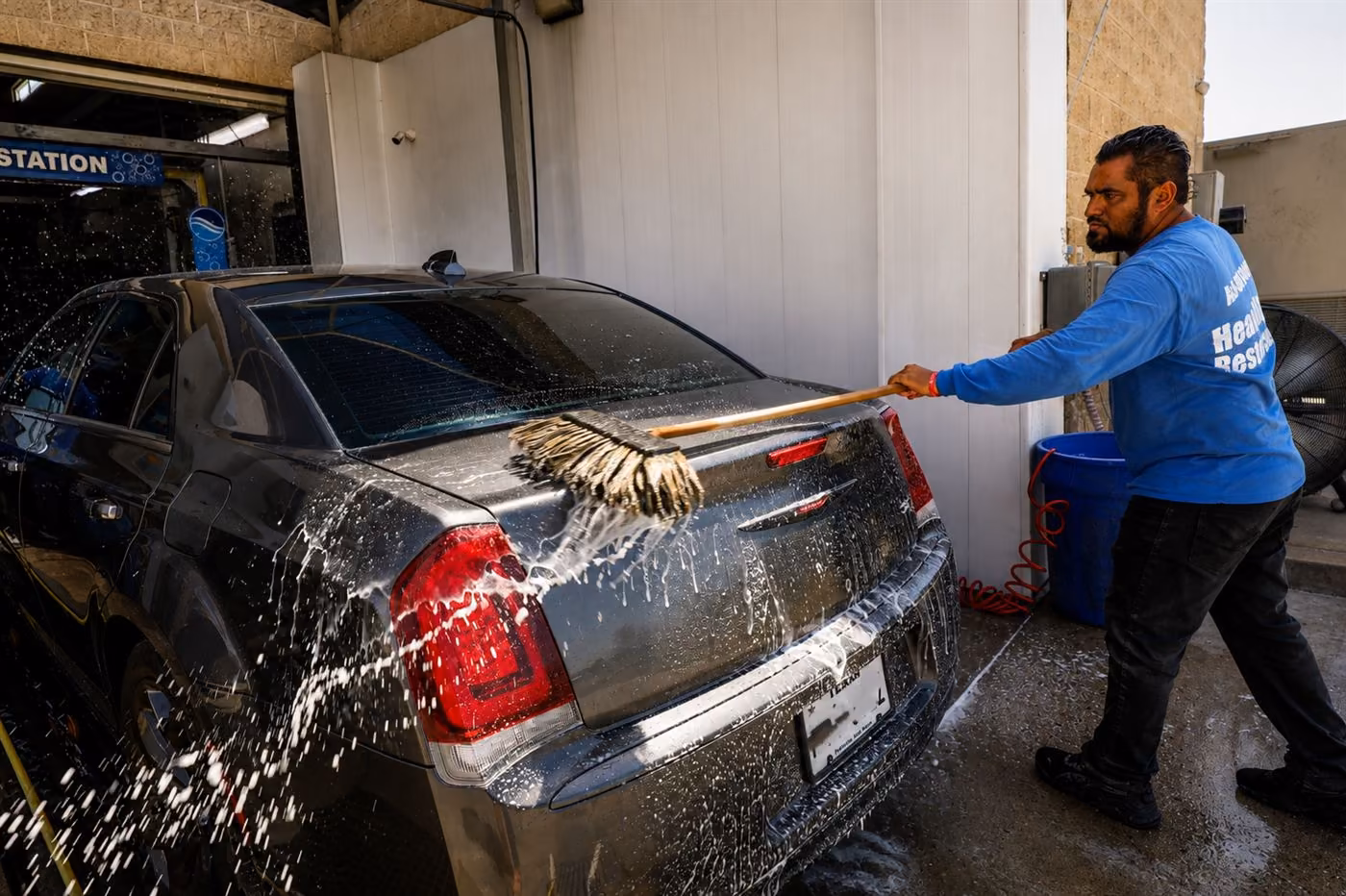 Crew member hand-prepping the rear of a dark sedan with a soft mitt as water sprays